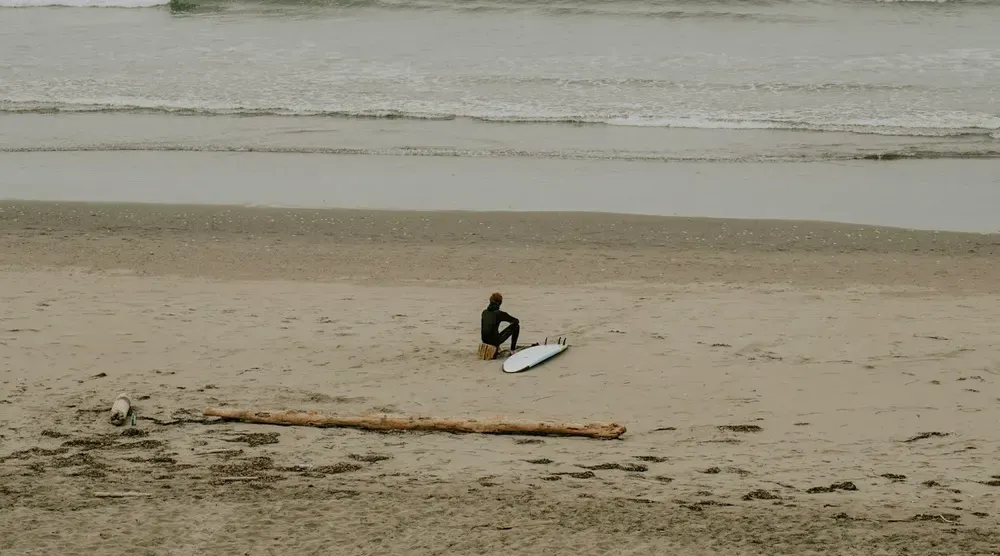 Surfeur assis sur le sable près de sa planche, face à l’océan